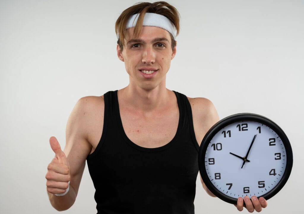 smiling young sporty guy wearing headband and wristband holding wall clock showing thumb up isolated on white background smiling young sporty guy wearing headband and wristband holding wall clock showing thumb up isolated on white background